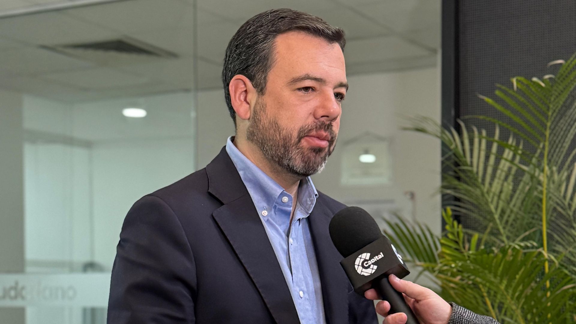 Hombre con traje negro y camisa azul hablándole a un micrófono