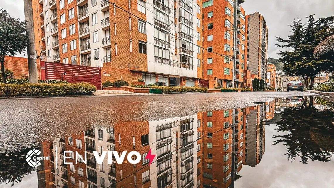 Edificios residenciales de ladrillo reflejados en un charco sobre calle mojada en Bogotá tras la lluvia, con entorno urbano y árboles