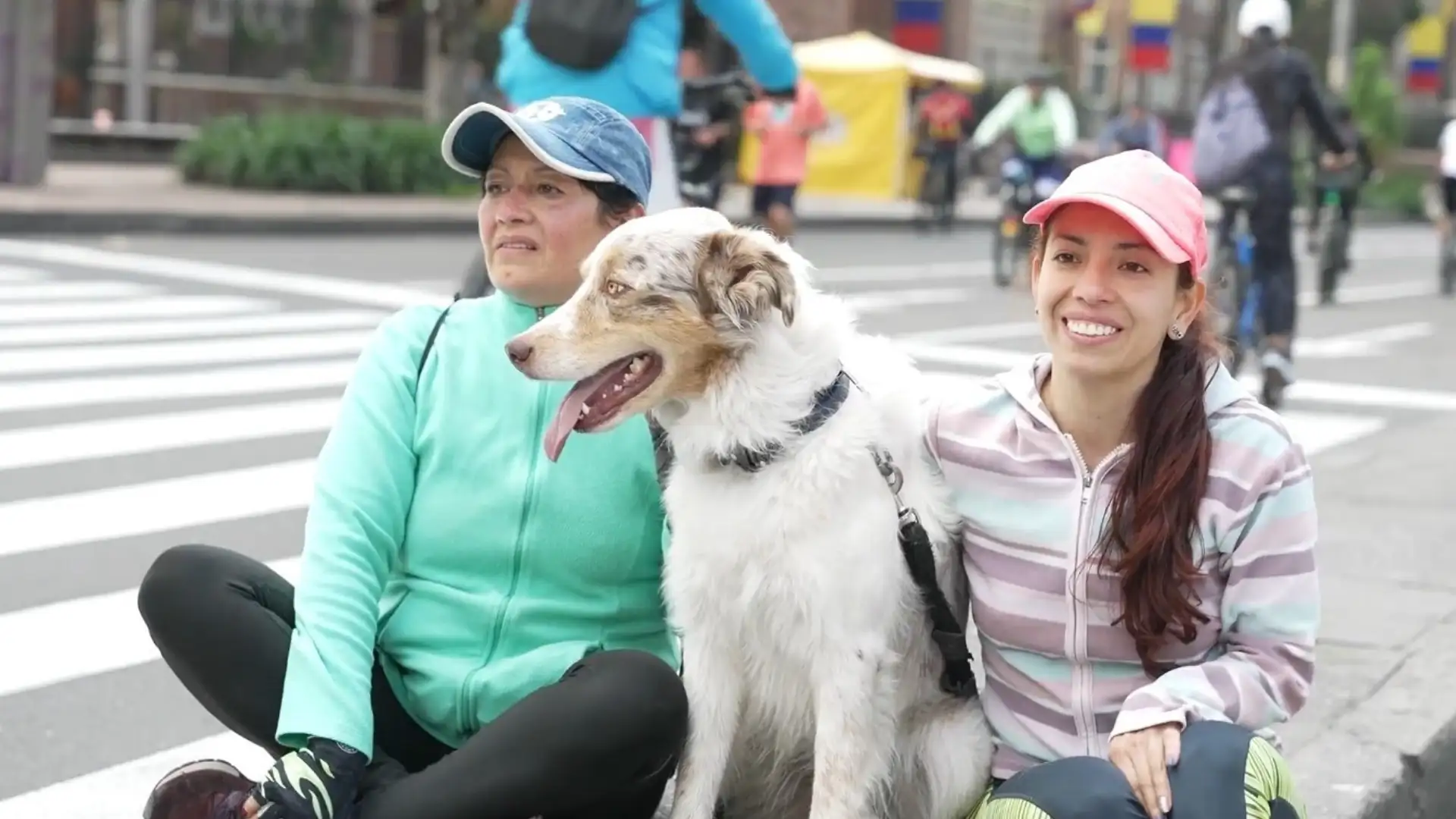 Foto tomada en la ciclovía en una calle de Bogotá en la que aparecen dos mujeres en ropa deportiva sentadas y en el centro un perro blanco con café y en el fondo se observan personas en bicicleta que van por la vía.