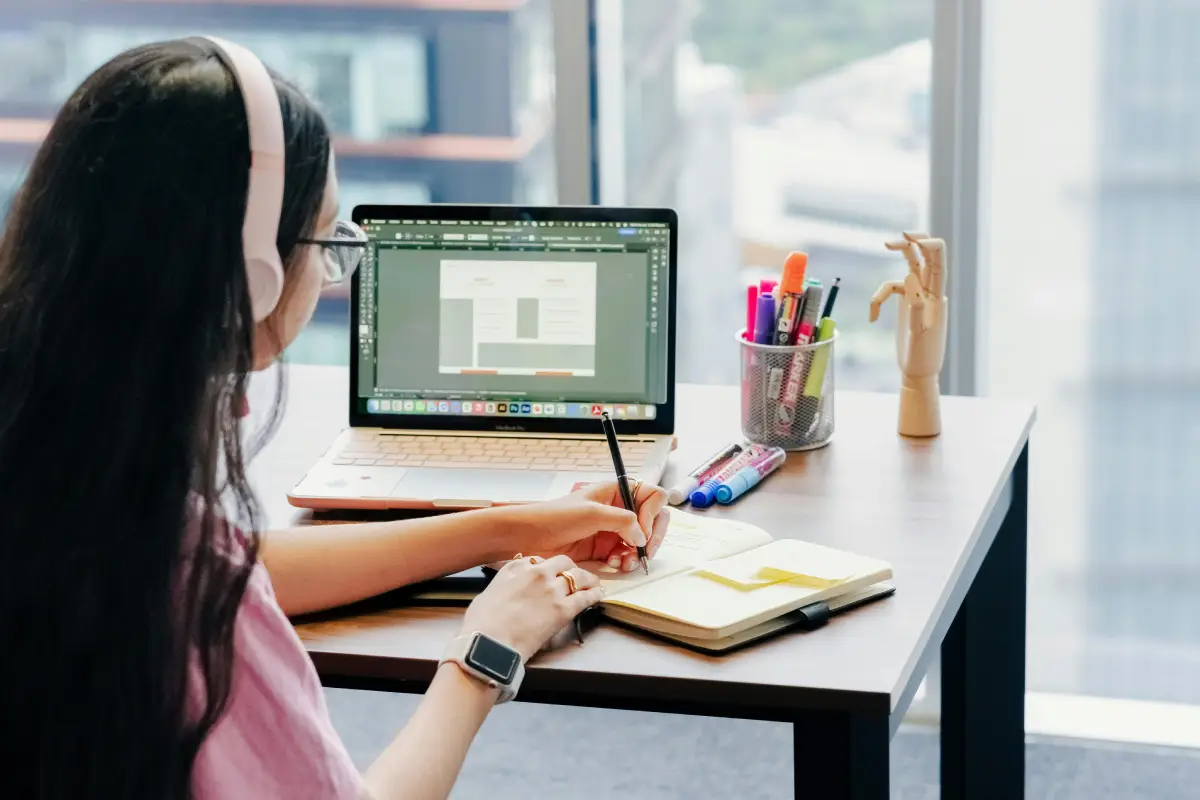 Una joven mujer escribe en su cuaderno mientras escucha música junto a su computador