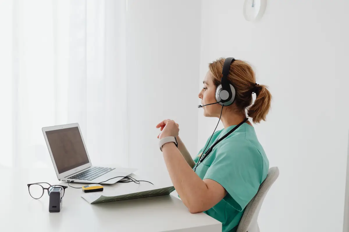 Una doctora usando un portátil para una consulta en línea, con auriculares en una oficina luminosa.