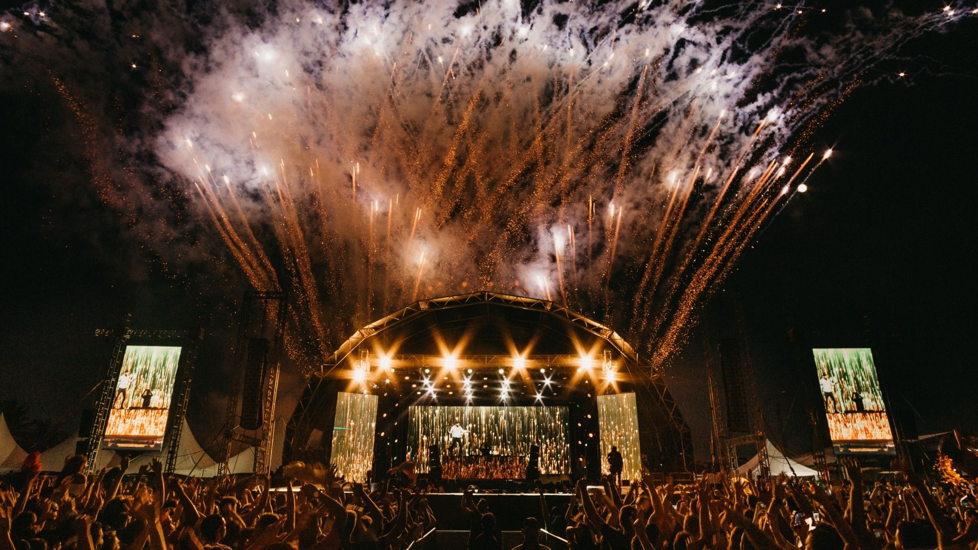 Concierto nocturno con luces brillantes en el escenario y un público entusiasta bajo el cielo estrellado.