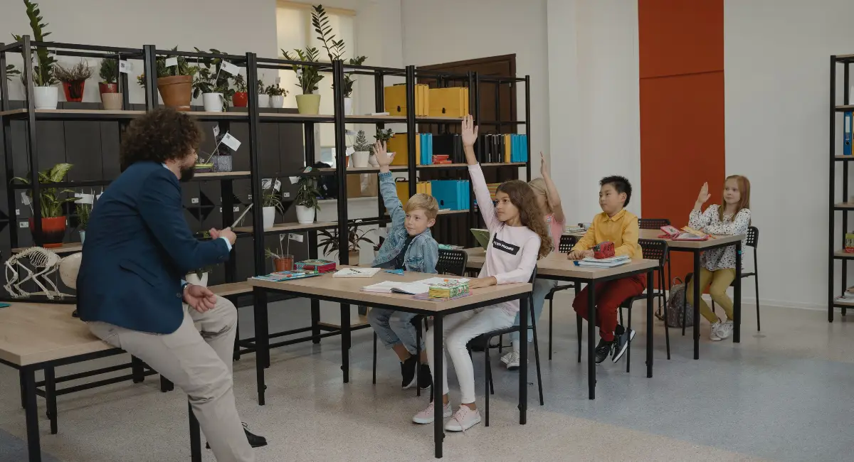Niños sentados en un aula de clase frente a un profesor alzando la mano para participar